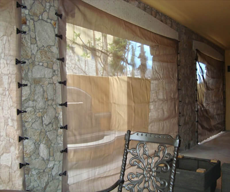 Transparent curtains, reminiscent of fabric hurricane protection for windows, hang over stone-framed windows, letting light into a patio area. In the foreground, a decorative metal chair and wooden box create a cozy, sheltered spot.