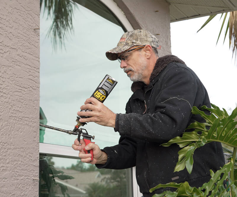 A man wearing a camo cap and glasses is applying sealant to a window frame outside a house with replacement windows. He is dressed in a dark jacket, holding a caulking gun. Greenery surrounds him, complementing the textured wall of the house.