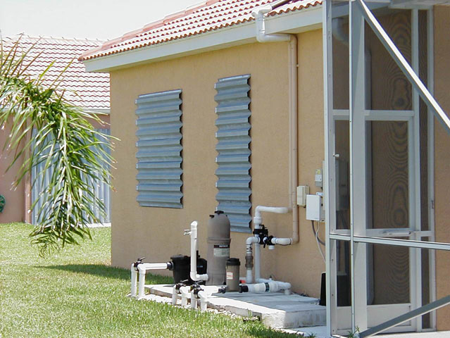 A beige house with a tiled roof features metal storm panels on its windows. In the foreground, pool equipment and pipes rest on a concrete pad, while a palm tree partially extends into the frame from the left.