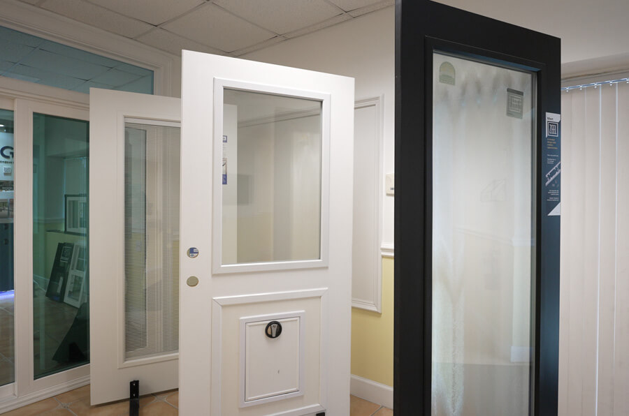 In a showroom brimming with possibilities, various door models are on display. A replacement white door with a large glass pane and mailbox slot captivates the foreground, while a black-framed door with frosted glass stands elegantly beside it.