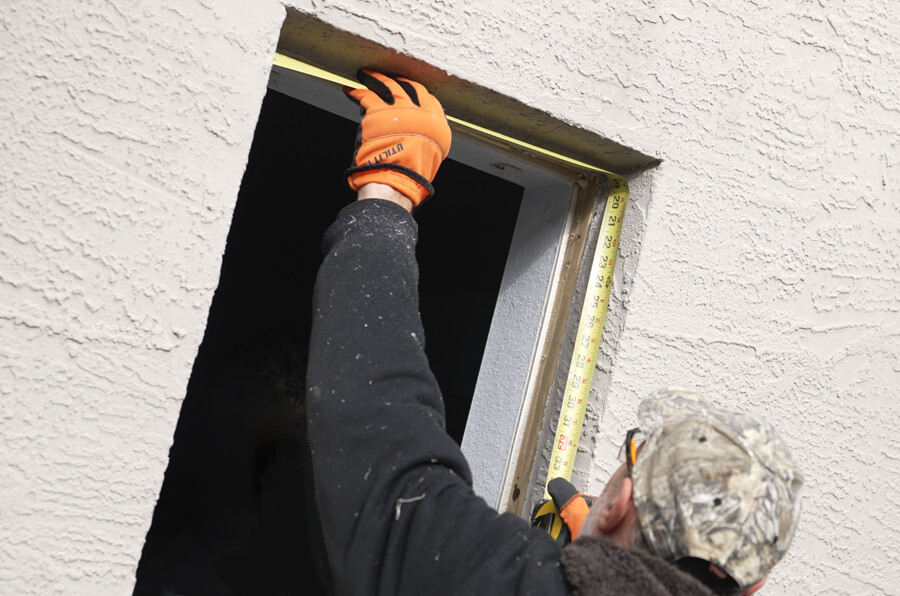 A person wearing orange gloves and a camo cap measures a window frame with a tape measure, preparing for a replacement window installation. They stand against a textured light-colored wall, extending the tape vertically to get the height of the frame.