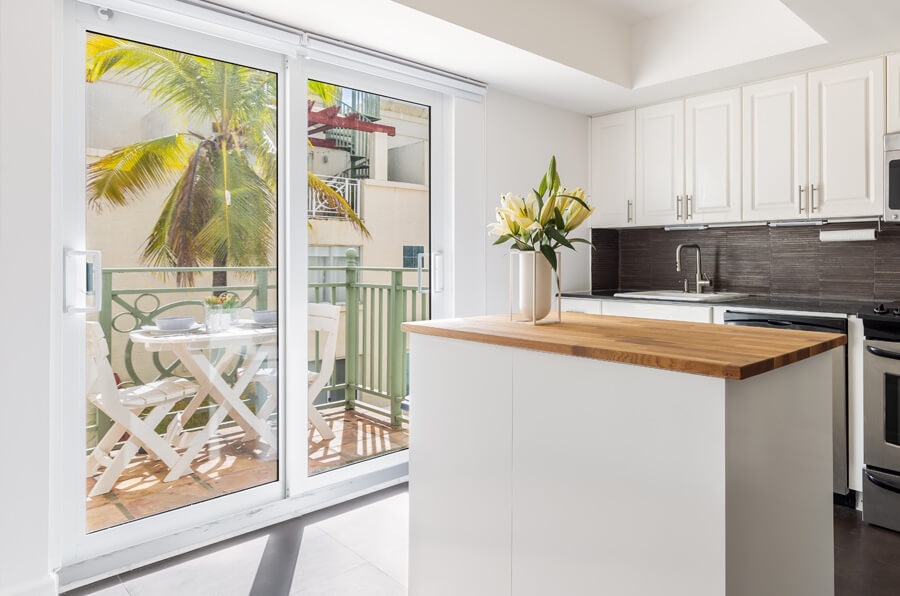 Bright kitchen with a wooden island, white cabinets, and stainless steel appliances. A vase with lilies sits on the island. Sliding patio doors open to a balcony with a table and chairs, enveloped by palm trees and sunlight.