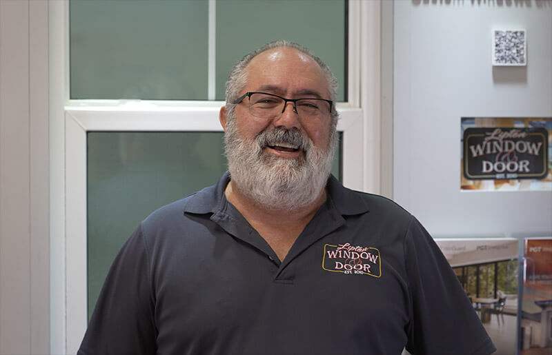 A smiling man with a gray beard and glasses stands in front of a window display. Tim Lipton, founder of Lipton, is wearing a dark polo shirt emblazoned with the logo Window and Door.